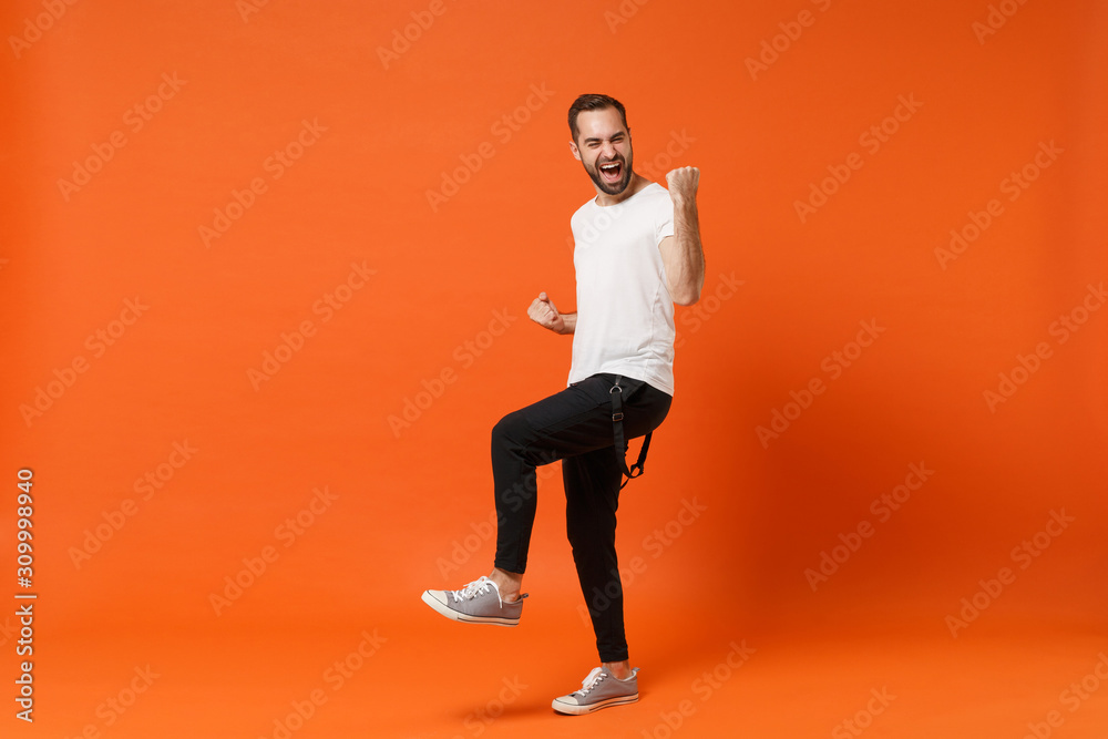 Overjoyed happy young man in casual white t-shirt posing isolated on bright orange wall background studio portrait. People sincere emotions lifestyle concept. Mock up copy space. Doing winner gesture.