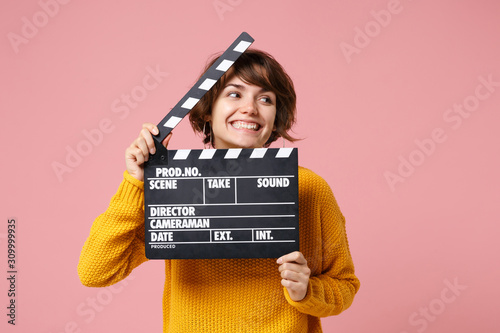 Cheerful young brunette woman girl in yellow sweater posing isolated on pastel pink background in studio. People lifestyle concept. Mock up copy space. Holding classic black film making clapperboard. © ViDi Studio