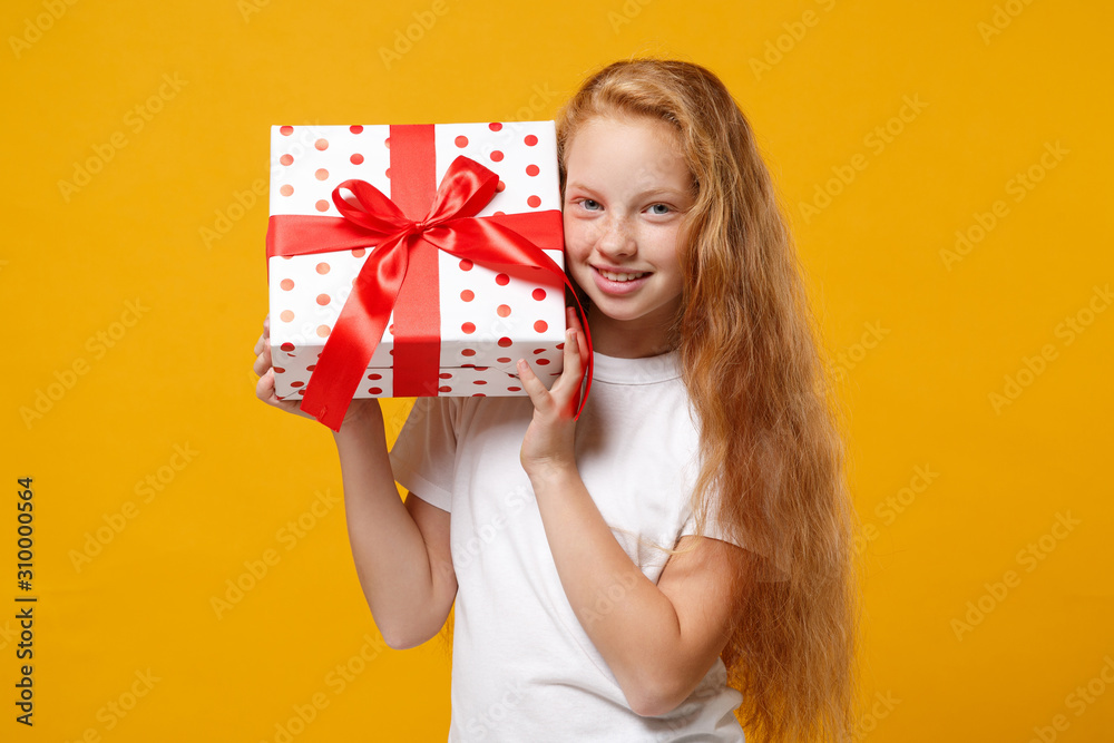 Smiling little ginger kid girl 12-13 years old in white t-shirt isolated on yellow background. New Year Women's Day birthday holiday concept. Mock up copy space. Hold present box with gift ribbon bow. Smiling little ginger kid girl 12-13 years old in white t-shirt isolated on yellow background. New Year Women's Day birthday holiday concept. Mock up copy space. Hold present box with gift ribbon bow.