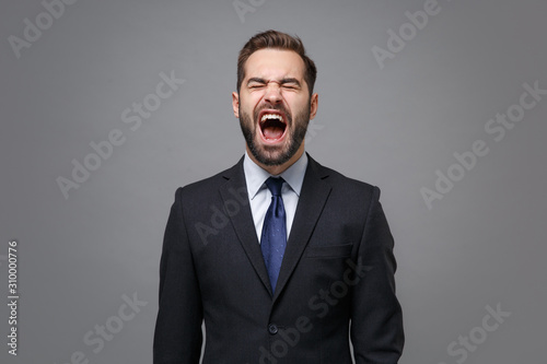 Frustrated young business man in classic black suit shirt tie posing isolated on grey wall background. Achievement career wealth business concept. Mock up copy space. Keeping eyes closed, screaming.