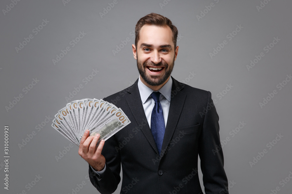 Cheerful young business man in classic suit shirt tie posing isolated on grey background. Achievement career wealth business concept. Mock up copy space. Holding fan of cash money in dollar banknotes. Cheerful young business man in classic suit shirt tie posing isolated on grey background. Achievement career wealth business concept. Mock up copy space. Holding fan of cash money in dollar banknotes.