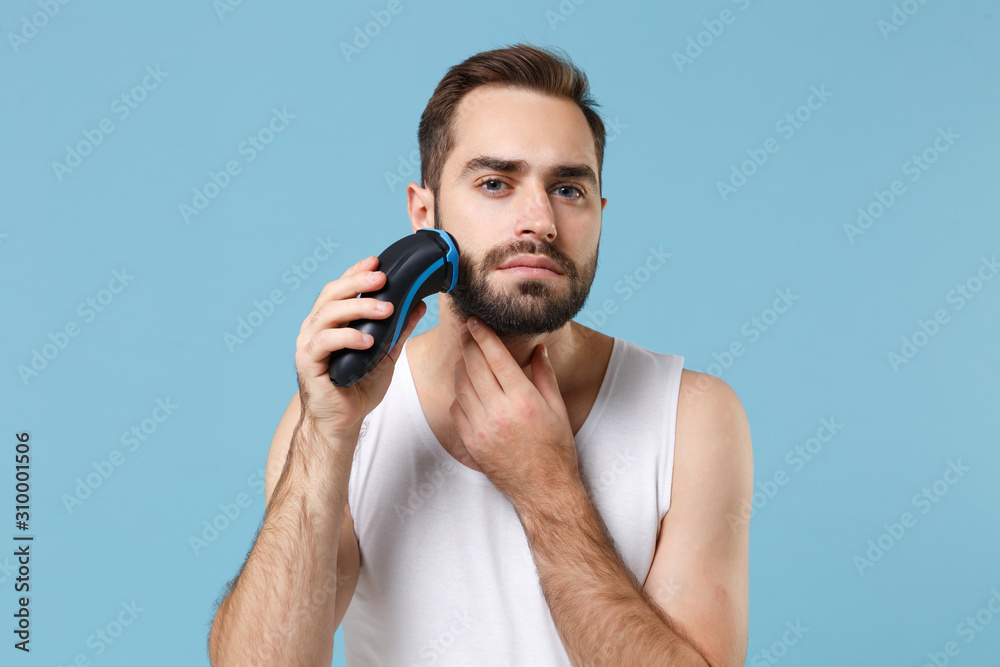 Naklejka premium Close up bearded young man 20s years old in white shirt hold electric razor isolated on blue pastel background, studio portrait. Skin care healthcare cosmetic procedures concept. Mock up copy space.