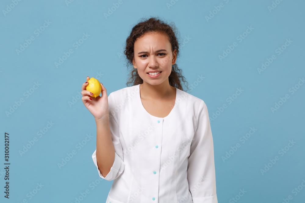 Displeased african american doctor woman isolated on blue background studio portrait. Female doctor in white medical gown hold lemon. Healthcare personnel medicine health concept. Mock up copy space. Displeased african american doctor woman isolated on blue background studio portrait. Female doctor in white medical gown hold lemon. Healthcare personnel medicine health concept. Mock up copy space.