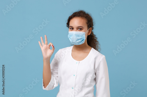 African american doctor woman isolated on blue background. Female doctor in white medical gown, sterile face mask showing OK gesture. Healthcare personnel health medicine concept. Mock up copy space.