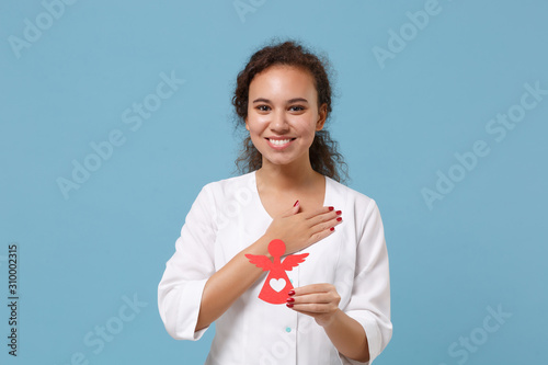 African american doctor woman isolated on blue background. Female doctor in white medical gown hold red toy angel put hand on chest. Healthcare personnel medicine health concept. Mock up copy space. photo