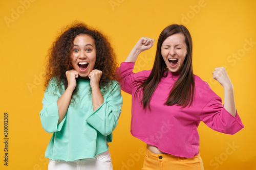 Two excited women friends european african american girls in pink green clothes posing isolated on yellow orange background. People lifestyle concept. Mock up copy space. Clenching fists like winner.