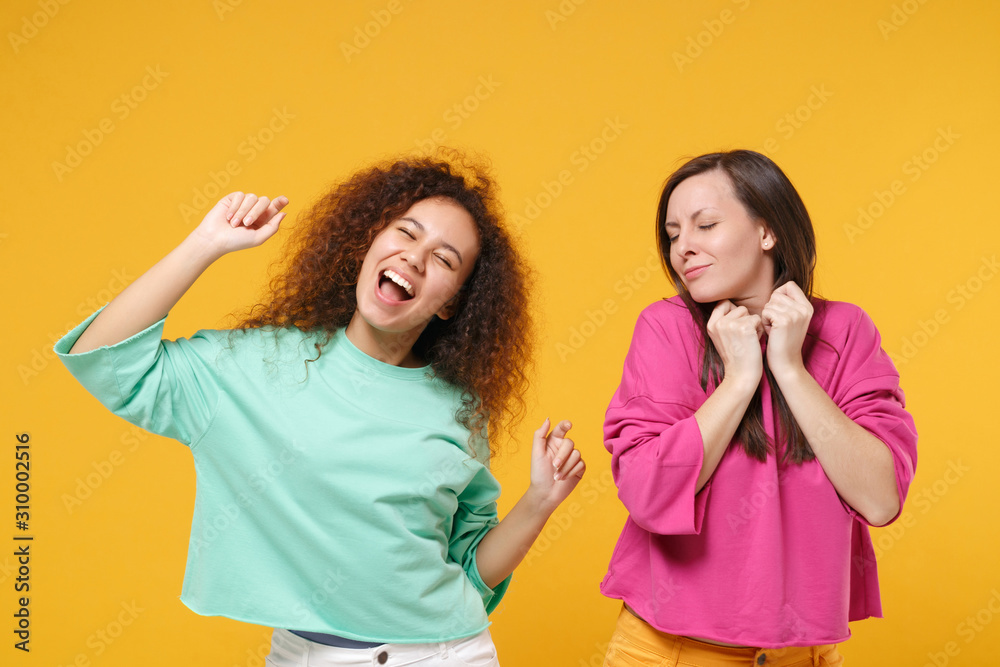 Two funny women friends european african american girls in pink green  clothes posing isolated on yellow background. People lifestyle concept.  Mock up copy space. Have fun dancing keeping eyes closed. Stock Photo |