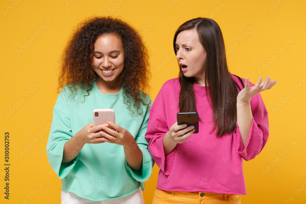 Two shocked women friends european african american girls in pink green clothes posing isolated on yellow background. People lifestyle concept. Mock up copy space. Using mobile phone, typing message. Two shocked women friends european african american girls in pink green clothes posing isolated on yellow background. People lifestyle concept. Mock up copy space. Using mobile phone, typing message.