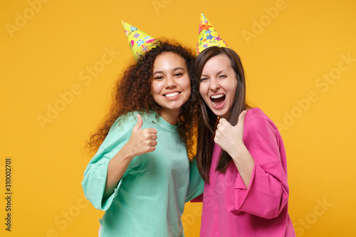 Two women friends european and african american girls in pink green clothes birthday hats posing isolated on yellow background. People lifestyle concept. Mock up copy space. Hugging showing thumbs up.