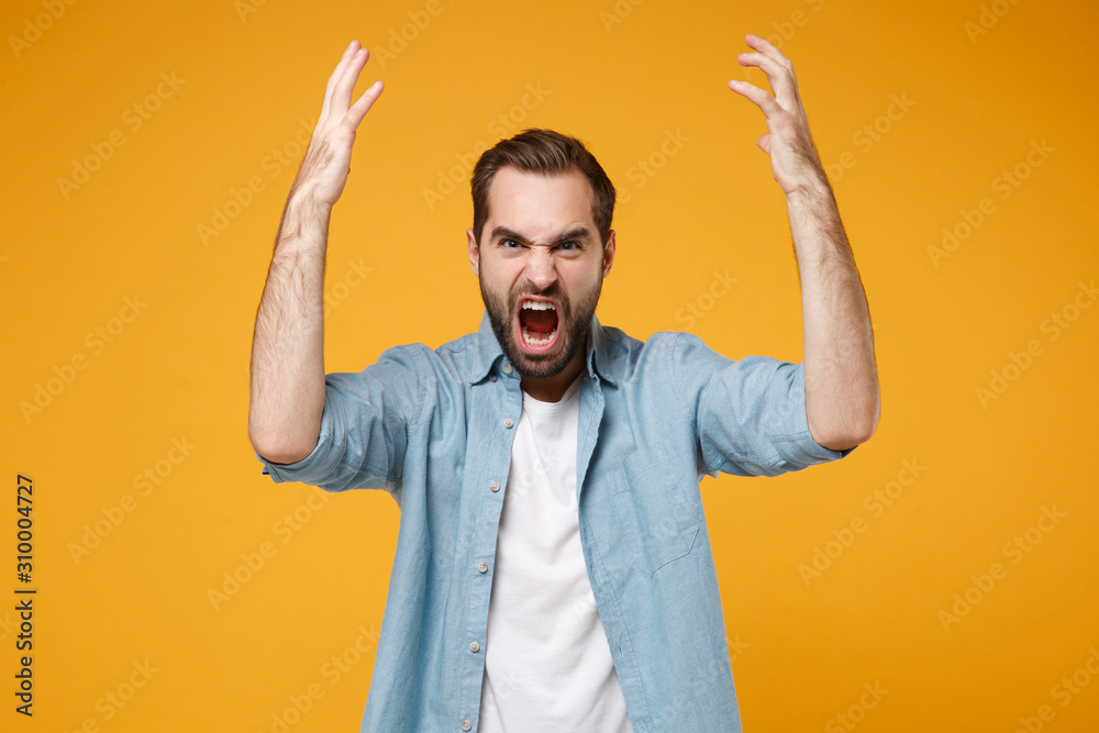 Fototapeta premium Angry young bearded man in casual blue shirt posing isolated on yellow orange wall background studio portrait. People emotions lifestyle concept. Mock up copy space. Screaming, spreading rising hands.
