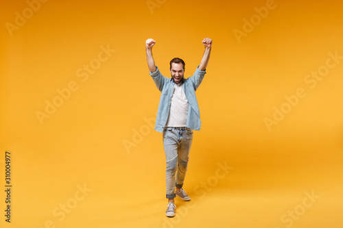 Joyful young bearded man in casual blue shirt posing isolated on yellow orange background studio portrait. People emotions lifestyle concept. Mock up copy space. Doing winner gesture rising hands up.