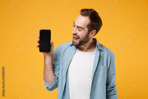 Cheerful young man in casual blue shirt posing isolated on yellow orange wall background, studio portrait. People lifestyle concept. Mock up copy space. Holding mobile phone with blank empty screen.