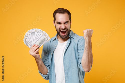 Crazy young man in blue shirt posing isolated on yellow orange background. People lifestyle concept. Mock up copy space. Holding fan of cash money in dollar banknotes, doing winner gesture, screaming. photo