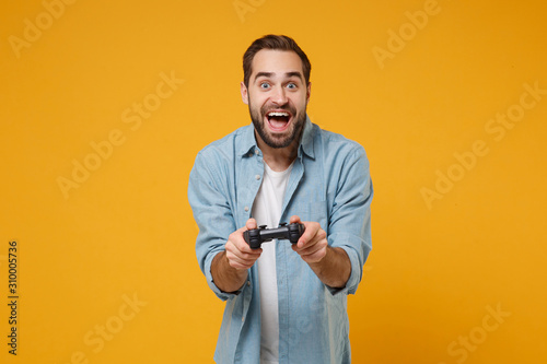 Excited young man in casual blue shirt posing isolated on yellow orange wall background, studio portrait. People sincere emotions lifestyle concept. Mock up copy space. Holding joystick playing game. © ViDi Studio