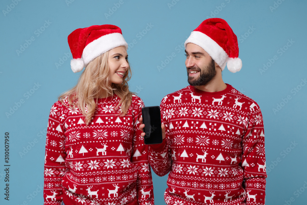 Smiling young couple guy girl in Christmas sweater Santa hat posing isolated on blue background. Happy New Year 2020 celebration concept. Mock up copy space. Hold mobile phone with empty blank screen. Smiling young couple guy girl in Christmas sweater Santa hat posing isolated on blue background. Happy New Year 2020 celebration concept. Mock up copy space. Hold mobile phone with empty blank screen.