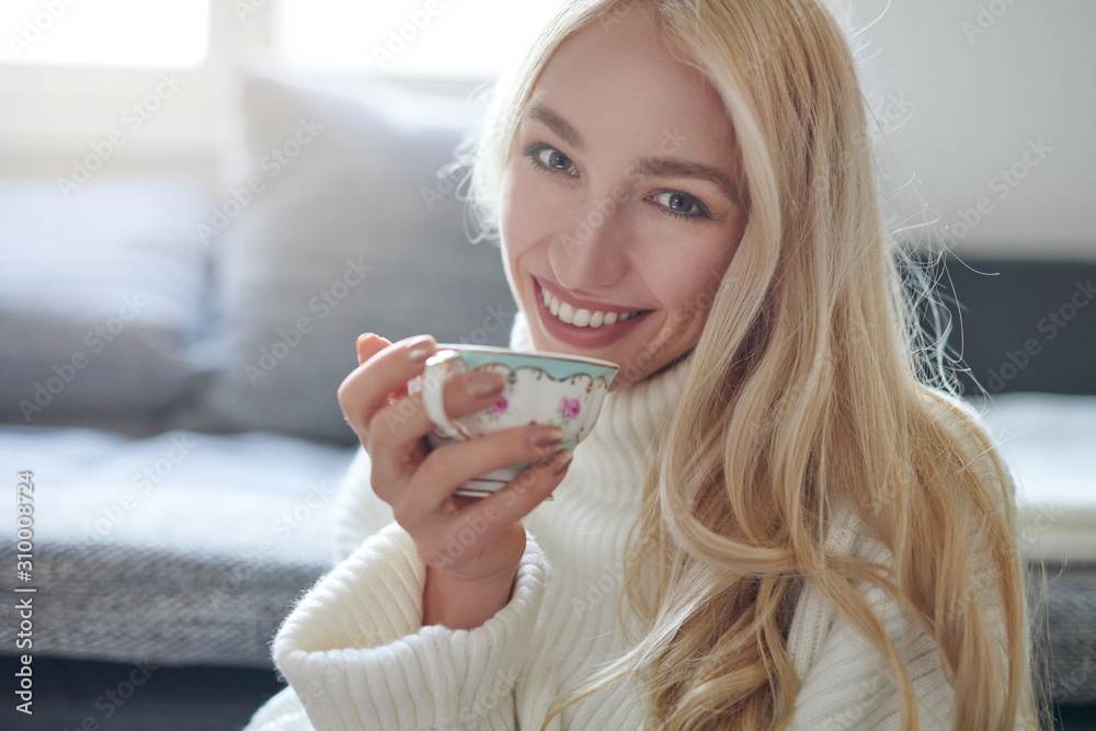 Pretty woman with long blond hair drinking tea from a porcelain cup looking at the camera with a sweet beaming smile of pleasure indoors at home Pretty woman with long blond hair drinking tea from a porcelain cup looking at the camera with a sweet beaming smile of pleasure indoors at home