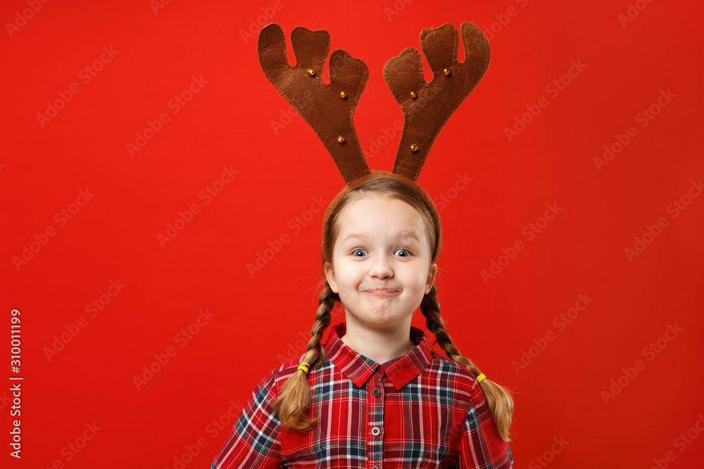 Funny happy child in red christmas dresse and deer horns on his head on a colored background. Emotional little girl in delight and surprise Funny happy child in red christmas dresse and deer horns on his head on a colored background. Emotional little girl in delight and surprise