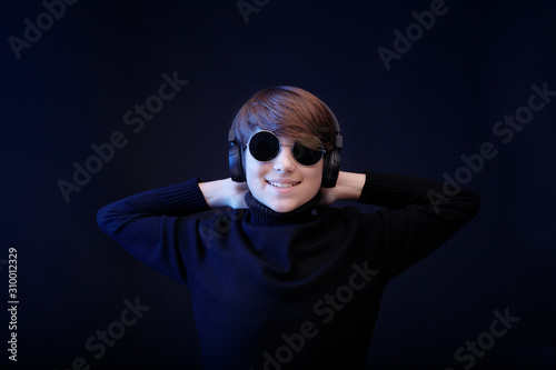Beautiful young boy in headphones listening music. Fashion portrait of teenager in glasses and stylish clothes. Black minimalism. Dark background.