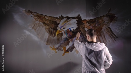 Artist designer draws an eagle on the wall. Craftsman decorator paints a picture with acrylic oil color. Painter painter dressed in a paint coat. Indoor. Dark magic cinematic look. © ivandanru