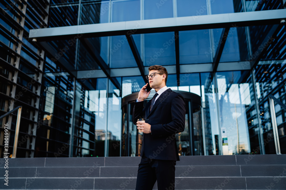 Fototapeta premium Trendy formal man speaking on mobile phone Young confident businessman in formal suit holding disposable cup of coffee while talking on smartphone standing against steps of office building