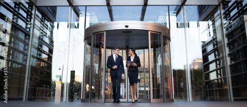 Smiling young coworkers talking on street
Contemporary elegant businesswoman with tablet talking to male colleague while walking out of modern office building with glass revolving doors
