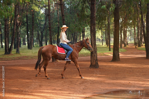 Young woman in shirt and straw hat, rides brown horse in the park