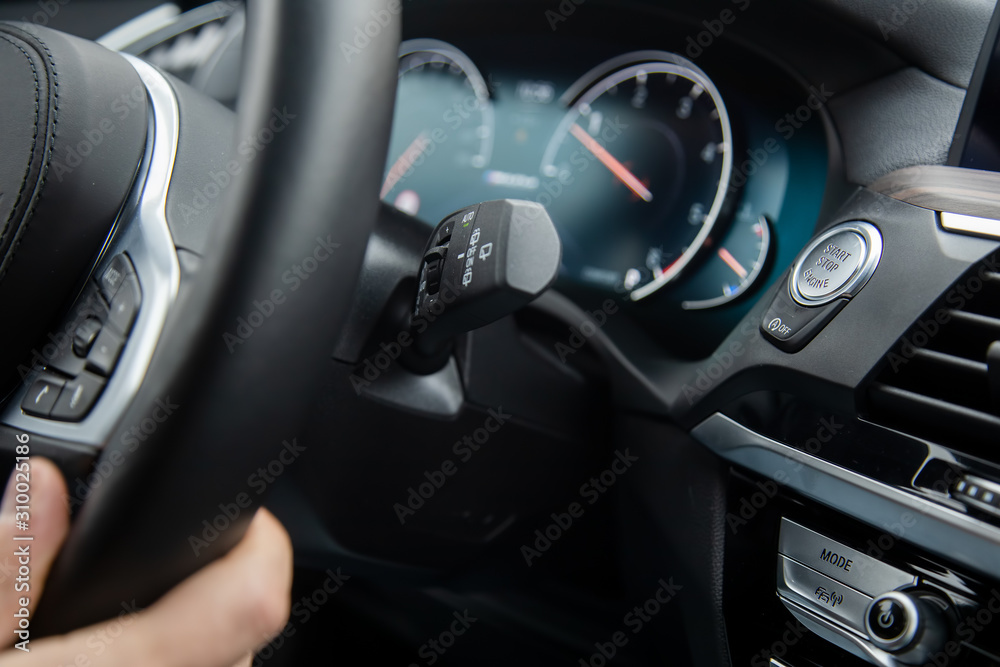 close-up start-stop button under the steering wheel of a modern car. man’s hand holds the steering wheel, lead nashina. speedometer in blur on the background