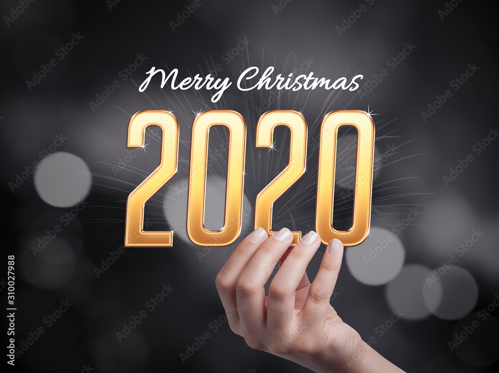 Female hand holding golden 2020 text and below of it merry christmas message in front of black and white background with bokeh effect. Female hand holding golden 2020 text and below of it merry christmas message in front of black and white background with bokeh effect.