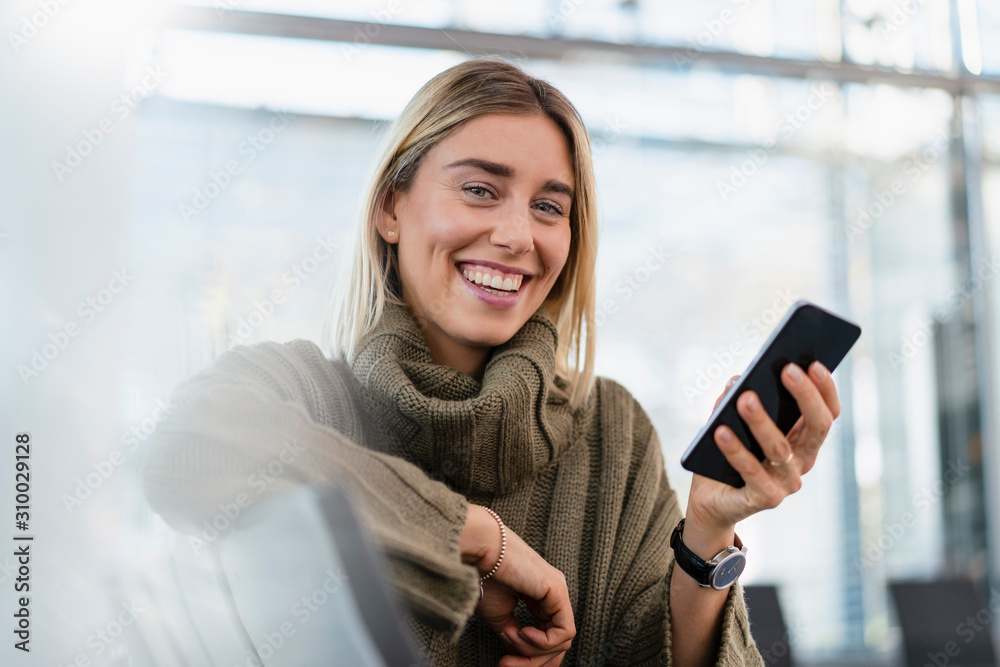 Portrait of happy young woman sitting in waiting area with cell phone