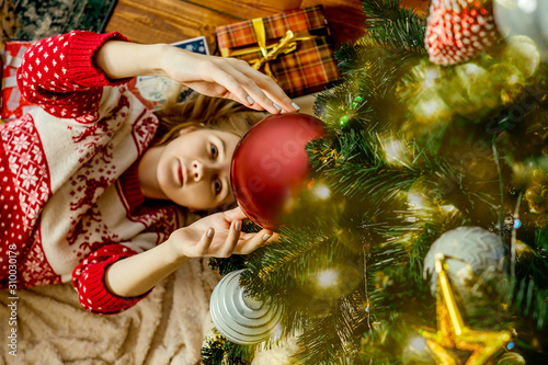 The girl lies under the Christmas tree and pulls her hands to the hanging big red ball. New Year's concept. Decorated Christmas tree with garland and lights.