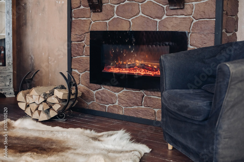 View over fireplace with burning logs, natural fur skin on the floor next to holder with logs in cozy room.