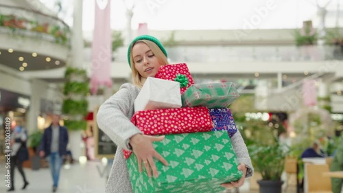Young attractive woman in hurry carrying a pile of colorful Christmas presents at shopping mall. Blonde girl in coat shopping gifts for family before boxing day. Consumerism.