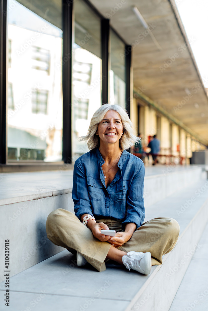 Mature businesswoman sitting cross-legged on steps in the city, using smartphone Mature businesswoman sitting cross-legged on steps in the city, using smartphone
