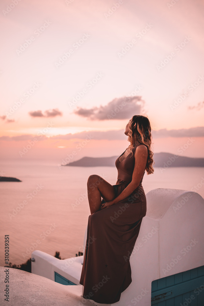 Young woman with blonde hair and purple dress watching the sunset in santorini greece - obrazy, fototapety, plakaty 