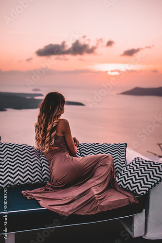 Fototapeta Naklejka Na Ścianę i Meble -  Young woman with blonde hair and purple dress watching the sunset in santorini greece