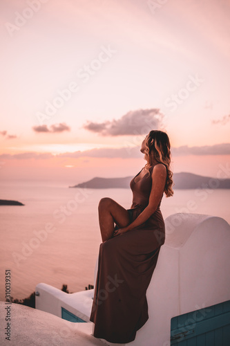 Fototapeta Naklejka Na Ścianę i Meble -  Young woman with blonde hair and purple dress watching the sunset in santorini greece