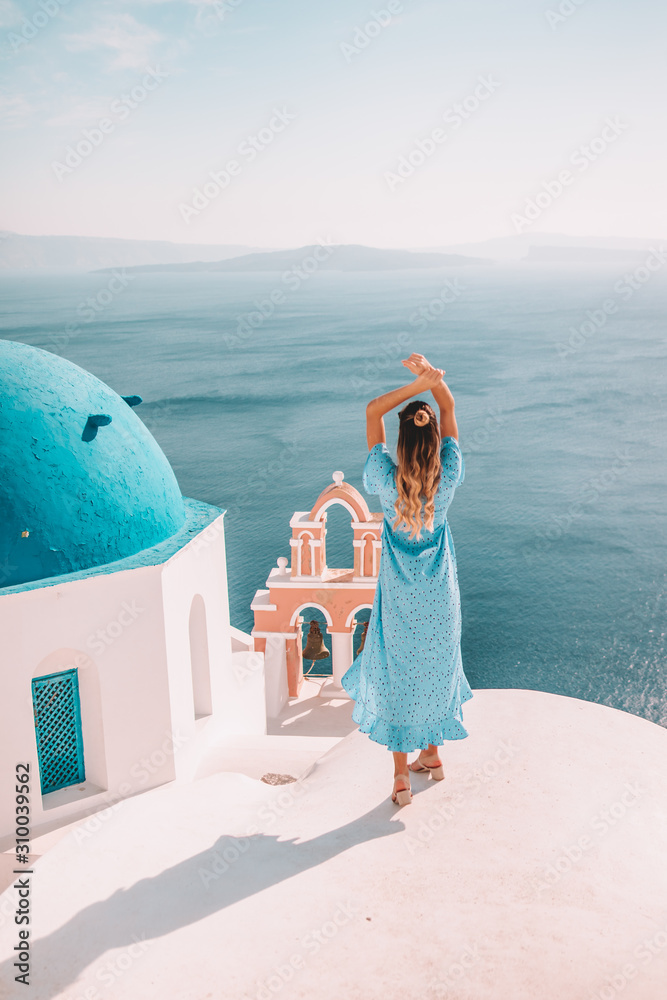 Young woman with blonde hair and blue dress in oia, santorini, greece with ocean view and churches - obrazy, fototapety, plakaty 