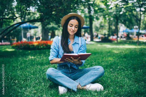 Attractive Ukrainian female student in fashionable hat sitting on green grass in city park and spending free time for literature hobby, positive Caucasian hipster girl in stylish outfit reading book