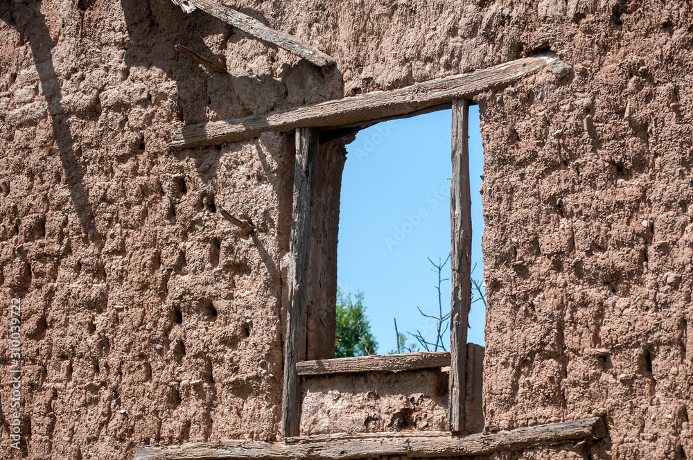 Brick wall with adobe clay plaster and broken wooden window frame remained of ruined old rural country house on cloudy blue sky background