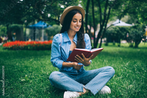 Happy cheerful hipster girl searching information in literature book during free time for reading popular literature book, smiling Caucasian female student learning with knowledge textbook in park