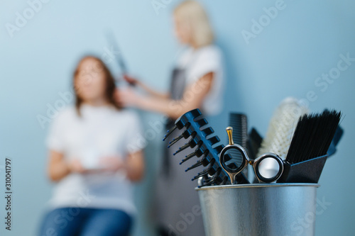 Barber tools in an iron bucket. Behind blurred scene of blonde female hairdresser in apron trimming client's hair over blue wall.