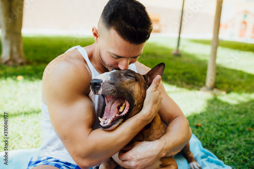 Muscular man sitting on blanket on a meadow hugging his dog