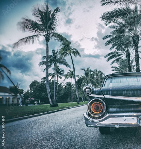Canvas Print Closeup of a vintage car on the road surrounded by palms and buildings under a c
