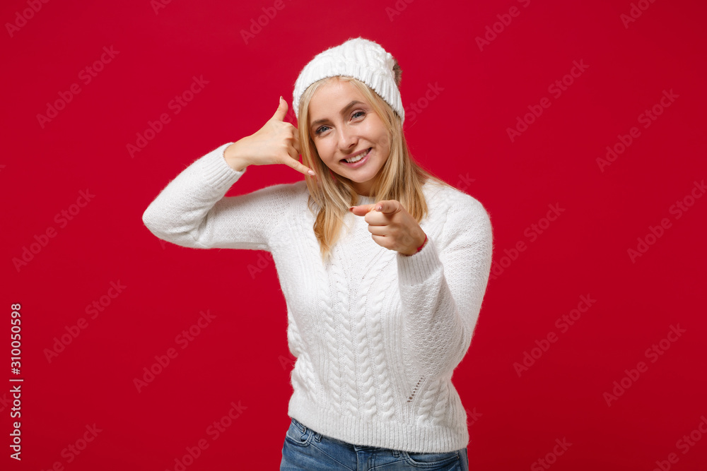 Smiling woman in sweater, hat isolated on red background. Healthy fashion lifestyle, cold season concept. Mock up copy space. Doing phone gesture like says call me back, point index finger on camera. Smiling woman in sweater, hat isolated on red background. Healthy fashion lifestyle, cold season concept. Mock up copy space. Doing phone gesture like says call me back, point index finger on camera.