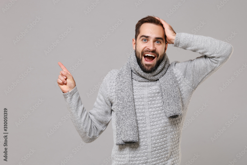Fototapeta premium Excited young man in gray sweater, scarf posing isolated on grey background in studio. Healthy fashion lifestyle cold season concept. Mock up copy space. Pointing index finger aside put hand on head.