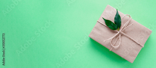 Gift box wrapped in kraft paper tied with twine decorated with two green leaves on neo mint background. Trend color 2020. Banner. DIY. Zero waste holidays concept. Flat lay. Copy space