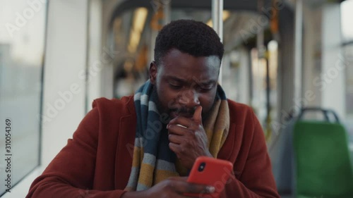 Sick poor handsome man sneezing allergy getting cold driving in tram to work. Modern young black businessman using smartphone reading news traveling in public transport.