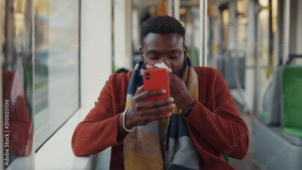 Bearded afro-american businessman using smartphone content suffering flu sneezing allergy with paper tissues driving to work in empty tram.