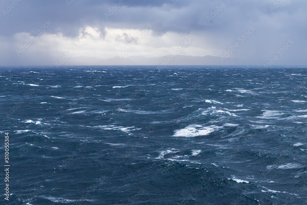 Grobe See im Atlantik, Europäisches Nordmeer vor Norwegen, mit Wellen und Gischt bei stürmischem Wind. Am Horizont dunkle Wolken mit Auflockerung, durch die die Küste zu sehen ist.
