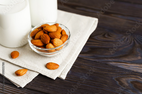 Close-up of almond milk in a glass mug and a refillable glass bottle, next to it lie almonds and a textile napkin on a wooden table.